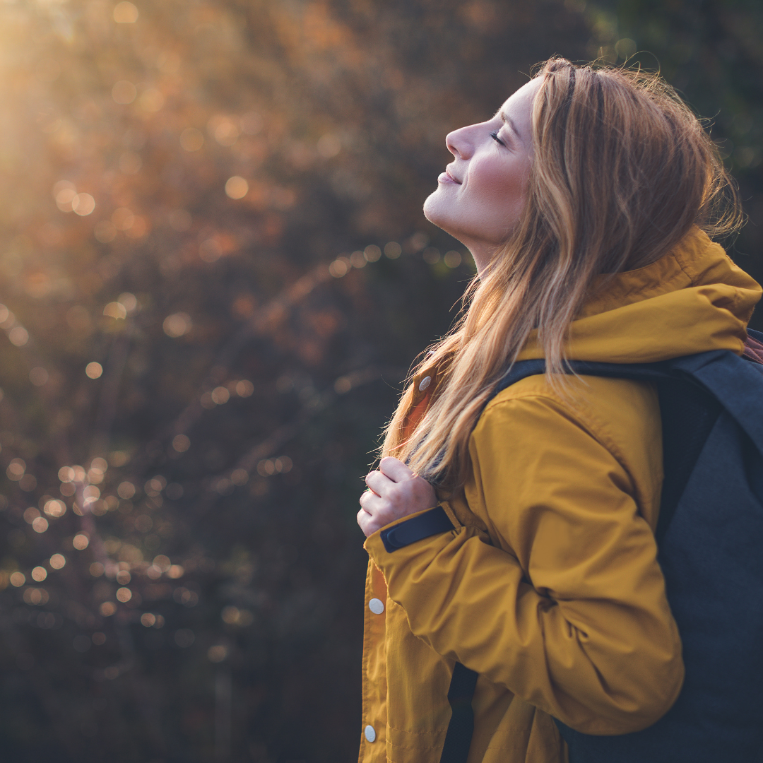 Une jeune femme dans la nature regarde en l'air. Elle porte un ciré jaune te un sac à dos noir.